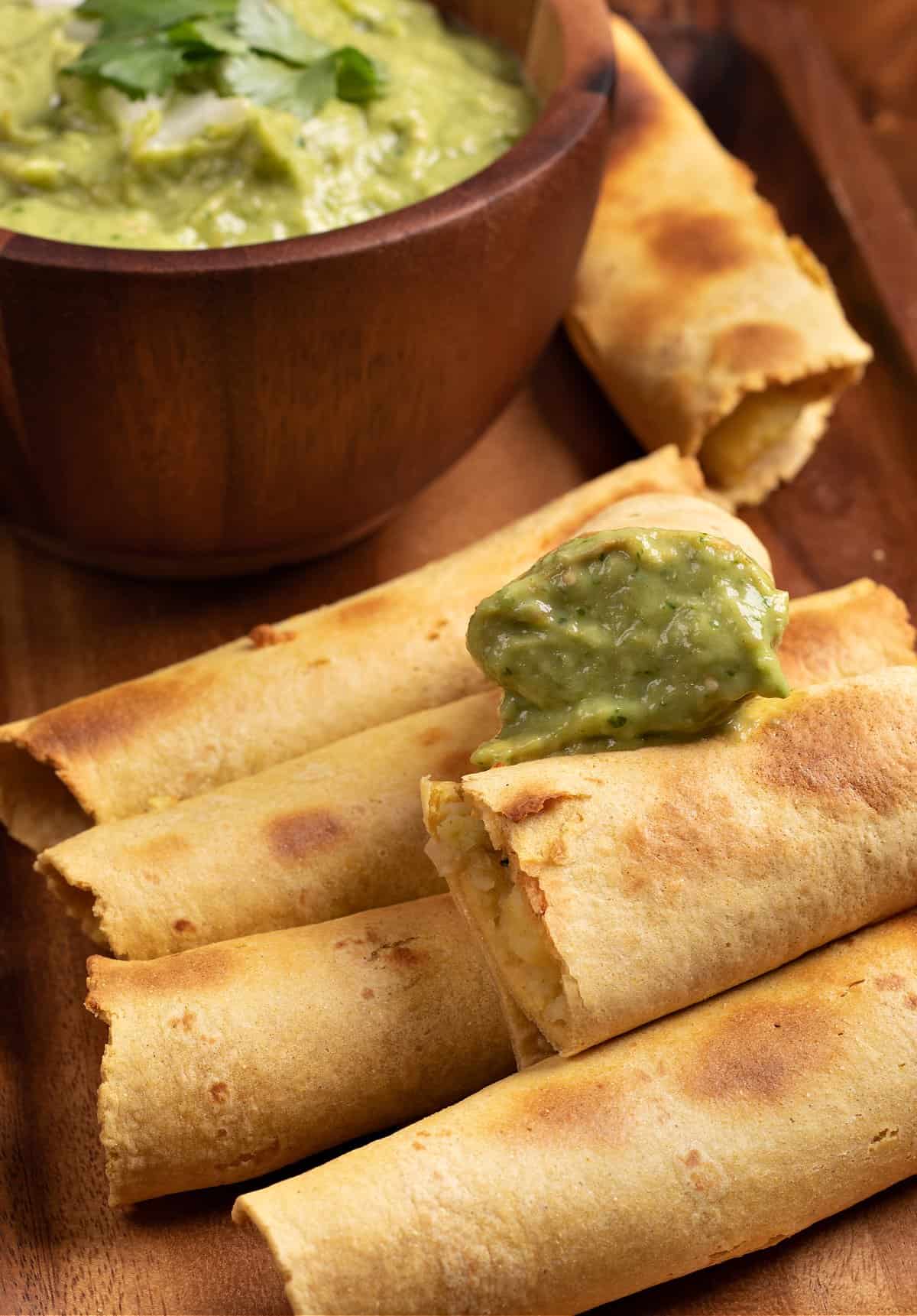 A tray of mashed potato taquitos with a bowl of tomatillo avocado salsa to dip in
