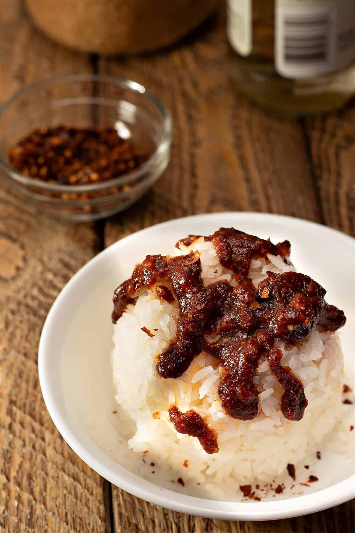 Bowl of rice drizzled with sweet chile sauce and red pepper flakes in the background