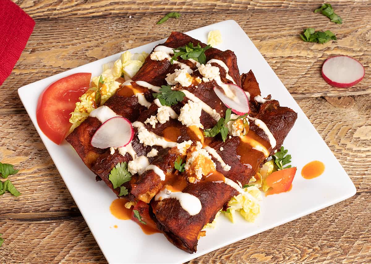 horizontal photo of 3 vegan enchiladas mineral sitting on a bed of cabbage slaw and topped with vegan queso fresco and vegan crema 