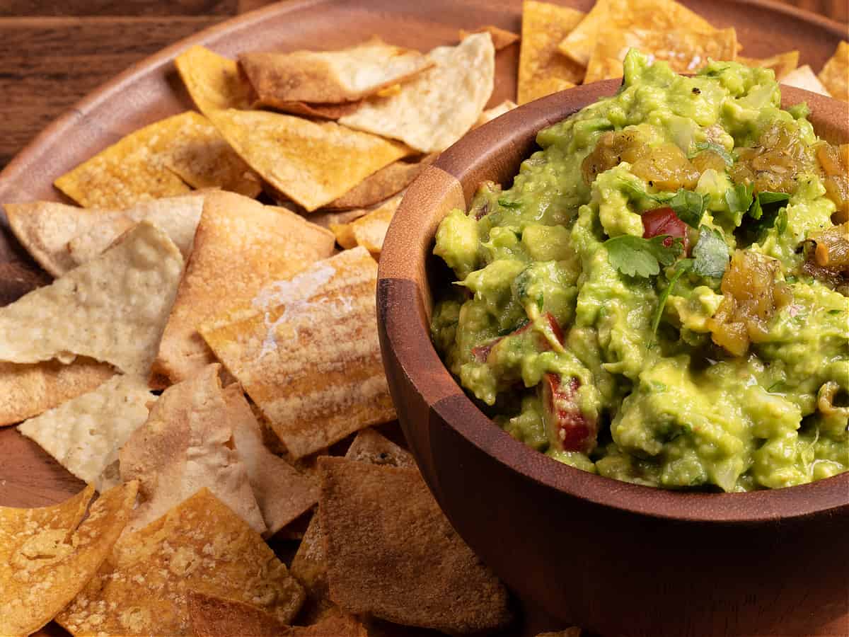 horizontal photo of a wooden platter of homemade tortilla chips and a bowl of vegan guacamole
